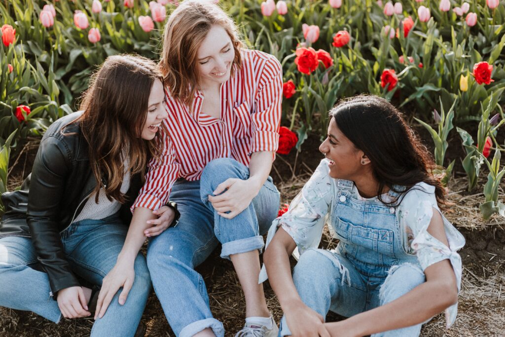 Three women sitting in a field of flowers while smiling and chatting. The woman in the centre has auburn hair and is wearing jeans and a red and white shirt