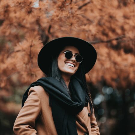 Woman in black hat, sunglasses and brown coat next to an Autumn tree with brown leaves.