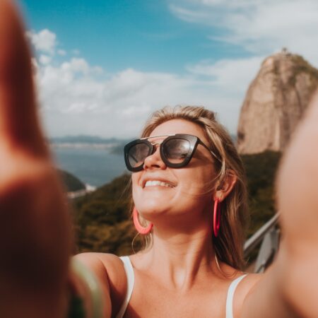 Blonde woman, in white vest top and sunglasses taking a selfie.