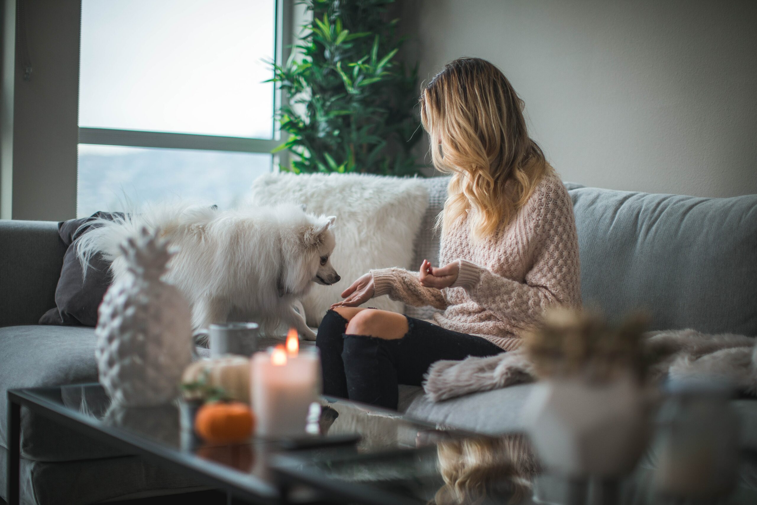 A woman with blonde hair, sitting on her cream sofa with her dog and some candles lit.