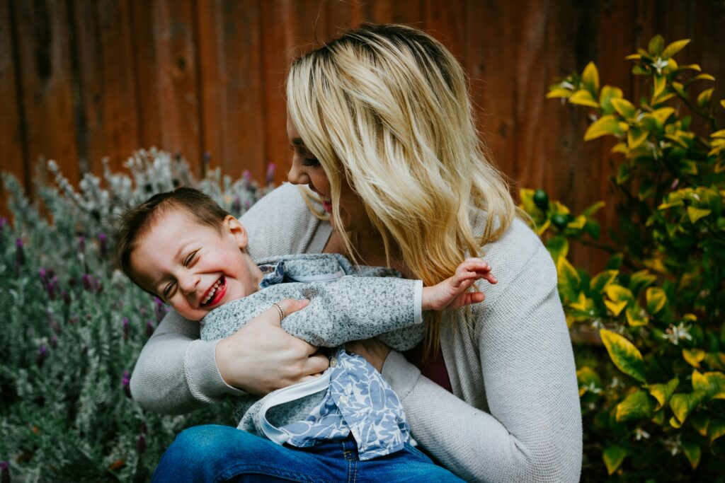 Woman with blonde hair in a shoulder length bob, wearing a a dove grey sweater, smiles and playfully cuddles her son with dark brown hair.