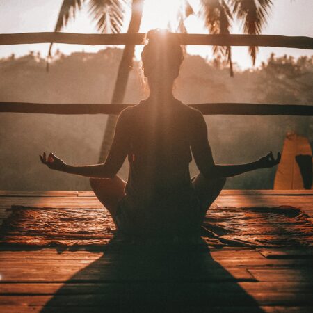 Silhouette of woman sitting cross legged in a yoga pose by a palm tree.