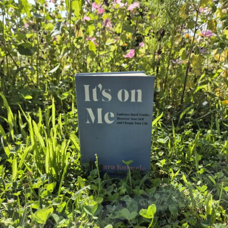 book called It's On Me, a blue cover with large white writing set against a green hedge with pink flowers in it.