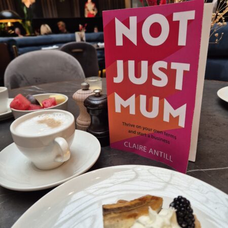 Not Just Mum with its bright pink cover and bold white font is placed on a cafe table, next to a Belgium waffle, complete with cream and fruit on a white plate, and a frothy cappuccino in a white cup.